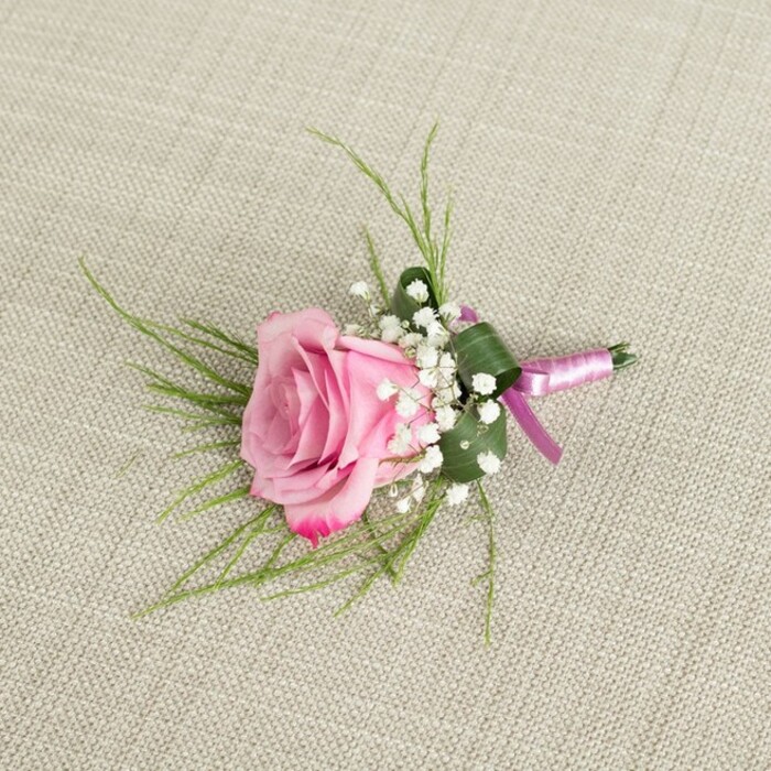 A finely crafted pink rose boutonniere is shown resting against a textured, light beige linen background, the woven threads of the fabric just visible enough to suggest a natural, understated elegance often favoured by couples marrying in and around Stanmore. At the heart of the design is a single rose in a rich yet gentle pink, its numerous petals tightly layered at the centre before loosening into soft, outward curls. The colour deepens almost imperceptibly towards the outer petal edges, where a slightly more vibrant magenta tone appears, echoing the romantic hues so popular for weddings and proms in this part of North London. Encircling the base of the rose, tiny white baby's breath flowers cluster together, their round, cloud-like blossoms adding airiness and a sense of classic formality. From behind the bloom, slender, bright green, grass-like strands of foliage fan out in different directions, creating a light frame that contrasts with the rose's solidity, while a broader dark green leaf is neatly folded and wrapped around the stems, acting as a natural collar that keeps the silhouette tidy and compact. The stems are carefully bound with a refined light pink satin ribbon, wrapped smoothly and tied into a small, elegant bow with short tails that lie flat against the linen. The ribbon's gentle sheen catches the soft, even light, as do the petals, which show subtle highlights and shadows that emphasise their velvety texture. This serene, close-up composition captures the quiet beauty of a handmade boutonniere, the sort that a groom, groomsman, or prom guest might have delivered from a trusted Stanmore florist for a celebration at a local hotel, synagogue, or family gathering space. It conveys thoughtfulness, intimacy, and the timeless charm of a single perfect rose worn close to the heart.
