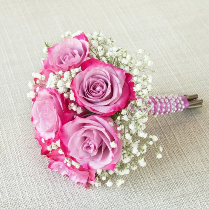 Glistening softly atop a light oatmeal-hued fabric-much like linens gracing tables at Stanmore's elegant gatherings-this compact, hand-tied bouquet beautifully balances modern flair with timeless charm. The focal point is a striking cluster of five roses, each exhibiting a masterfully blended gradient: the heart of each bloom is a tranquil lavender, gently radiating outward to brilliant, deep pink and magenta fringes. Dancing around the roses, sprays of white baby's breath (gypsophila) lend an ethereal, frothy texture, catching and diffusing the light to enhance the bouquet's airy, sophisticated mood. The stems are bound in layers of rich purple satin ribbon, spiraling seamlessly down to a finish adorned with lustrous rows of white pearls, each one reflecting soft, natural light. The natural tips of green stems emerge below, speaking to the freshness and authenticity of locally sourced blooms-perhaps gathered early morning from growers near Bernays Gardens. Seen from above, the bouquet's rounded, balanced shape and intricate accents are revealed, making it an inspiring centerpiece for Stanmore weddings, engagement parties, or intimate dinners at home. This bouquet from Flowers Stanmore perfectly captures the town's enduring love for quality, blending subtle luxury, expert floristry, and heartfelt sentiment in every detail.