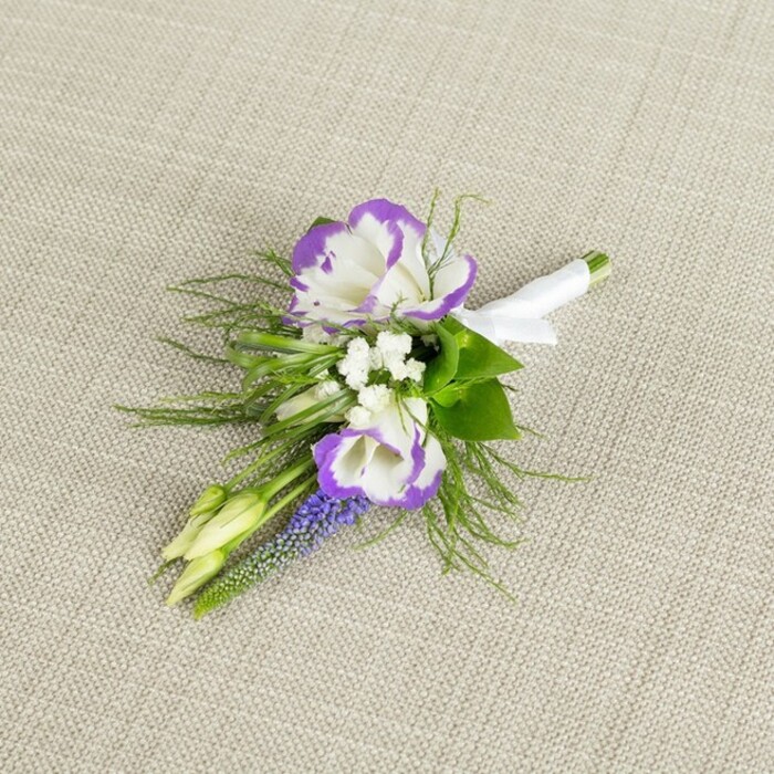 Set against a softly textured, light beige fabric backdrop, this boutonniere is captured in a close, detailed view that highlights both craftsmanship and natural beauty, as if laid out in a quiet Stanmore bedroom just before dressing for the day. Dominating the small arrangement are two lush lisianthus blooms with silky white petals edged in a bold, regal purple, the ruffled margins creating a sense of movement and depth. Tucked between and around these main flowers are dainty sprays of tiny white filler blossoms, much like baby's breath, that lend a Romantic softness and a hint of vintage wedding charm. Standing upright through the arrangement, a narrow spike of deep lavender-purple muscari, or grape hyacinth, offers a distinctive texture: tiny, bead-like bells packed tightly together, echoing the lisianthus colouring with a moodier hue. Surrounding the flowers, fine, feathery greenery reminiscent of asparagus fern drapes delicately, its light fronds contrasting with a few slightly broader, darker green leaves that ground the composition and suggest fresh garden foliage. Two pale green buds remain closed, symbolising new beginnings and lending a sense of living freshness, just as you might find in early spring along the paths toward Canons Park. At the base, all the stems are precisely aligned and wrapped in a smooth white satin ribbon, carefully wound and secured in a small, tidy bow, clearly ready to be pinned onto a lapel without fuss. The palette of white, purple, and green conveys quiet sophistication and romance, making this boutonniere perfect for a groom or groomsman attending an elegant Stanmore wedding or a formal event in one of the area's graceful venues.