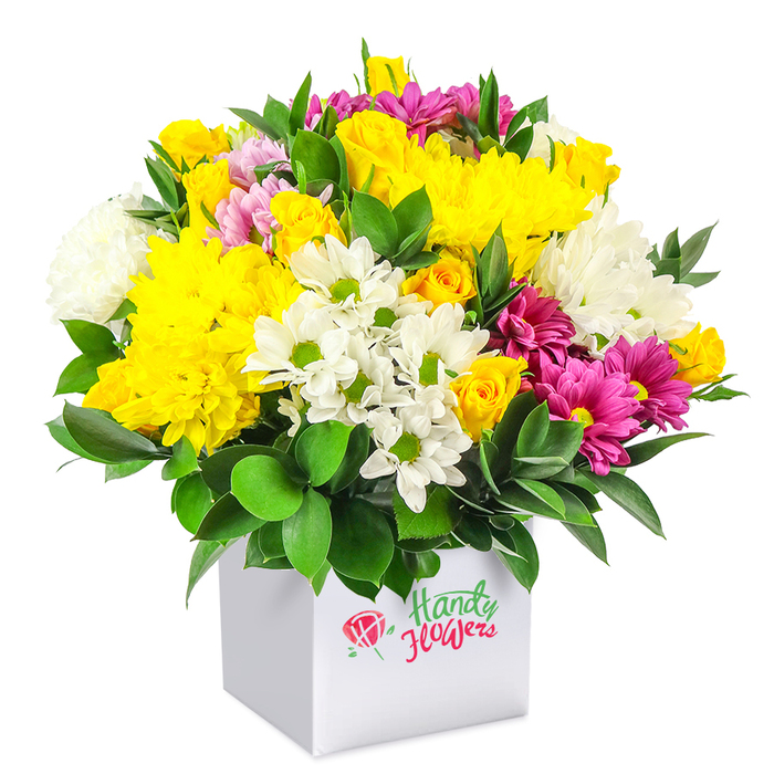 A richly textured mixed-flower arrangement in shades of yellow, white, and fuchsia sits in a sleek white square box, photographed against a bright white backdrop, as if styled in a contemporary Stanmore living room. The bouquet forms a smooth, rounded dome, every space between blossoms carefully filled to create a sense of fullness and generosity. Yellow chrysanthemums take centre stage, their densely layered petals forming plush blooms that feel almost cushion-like to the eye. Around them, yellow spray roses cluster in small groups, some fully budded, others just beginning to unfurl, giving the design a sense of life and gentle movement. White daisy-like chrysanthemums, each face marked by a fresh green centre, thread through the warmer colours, bringing lightness and a touch of countryside simplicity, while larger white chrysanthemums contribute body and a soothing, cool contrast. Punctuating this pastel balance are vivid fuchsia and deep magenta chrysanthemums, their saturated tones adding flashes of excitement, a nod to the vibrant community atmosphere around Stanmore Broadway and local cafés. Dark green foliage frames the entire arrangement, with leaves that appear smooth and glossy, providing visual depth and helping to define the bouquet's rounded silhouette. The white cube box is understated and modern, its clean edges and matte finish allowing the flowers to command attention, much like a carefully chosen accent piece in a Stanmore home. The lighting is clear and even, highlighting fine details such as the subtle curves of each petal and the healthy sheen on the leaves. Overall, the image conveys a joyful, thoughtfully composed floral gift, perfectly suited for birthdays, thank-you messages, housewarmings, or everyday celebrations delivered across Stanmore and nearby neighbourhoods.