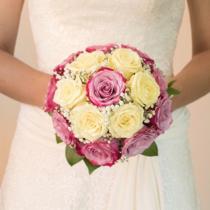 Captured at the moment anticipation blooms, a bride in Stanmore displays her exquisite rounded bouquet, inspired by the area's graceful gardens and classic traditions. The bouquet is an immersive composition of tightly packed lemon creme roses at its heart, encircled by dramatic fuchsia pink and gentle lavender-blushed roses, their colours playfully shifting under the soft, even light. Bursts of tiny white Gypsophila weave among the petals, echoing the lightness of a spring breeze along Stanmore Hill. A few rich green leaves edge the bouquet, providing an organic, grounded accent. Held patiently at waist level, the bouquet is flanked on either side by the bride's bare arms, her white dress featuring decorative lacework that mirrors the intricacy of the floral arrangement-a nod to the timeless style seen during elegant events in the Stanmore area. The lower skirt flows smoothly, catching a subtle gleam from the daylight. The overall ambiance evokes a sense of calm joy found in a carefully orchestrated celebration, where every element-from blush of petal to swirl of lace-speaks of local craftsmanship, artistry, and the dreams that gather for weddings in Stanmore.