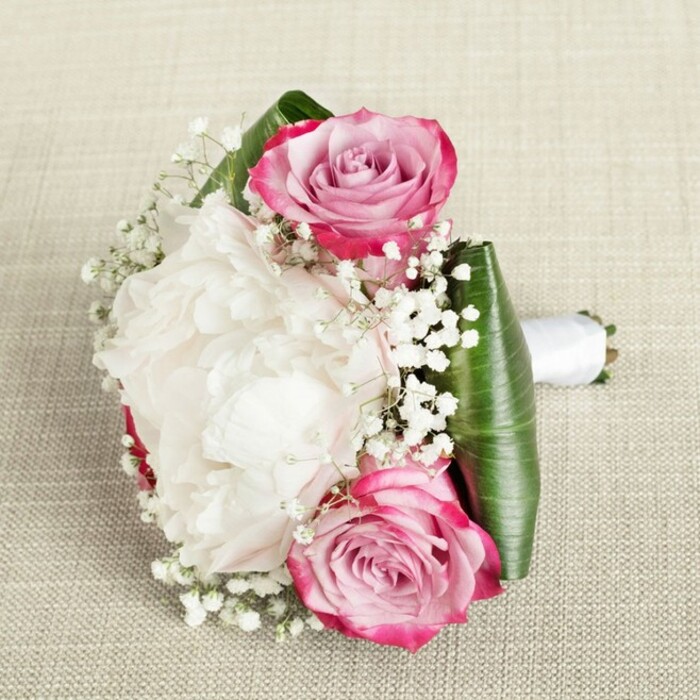 From an elegant, slightly raised viewpoint on a beige linen canvas evocative of Stanmore's graceful estates, this compact bouquet brings together luxury, romance, and expert floristry. The central feature is a breathtaking open white peony-its soft, ruffled, and voluminous petals forming a plush, cloud-like centrepiece. On either side, two pink roses bring dynamism to the arrangement, their outer edges rich in magenta and fuchsia, blending seamlessly into soft blush and pale lavender toward the intricately coiled core. These blooms, fresh and robust, recall the classical gardens that dot Stanmore's landscape. Throughout, delicate baby's breath infuses the design with lightness and an almost celestial airiness, filling gaps with a playful, whimsical texture. Bold, lustrous green leaves, expertly folded, wrap around the base providing a structured and contemporary finish, while a smooth white satin ribbon secures the stems, creating a poised and comfortable handle. Bathed in gentle, natural light, the bouquet's textures and colour gradients come alive, offering a timeless visual that would be perfectly at home in Stanmore's charming wedding venues or among the manicured lawns of Bentley Priory. Every element is a testament to the thoughtful selection and arrangement offered by the craftsmen at Flowers Stanmore, delivering beauty and elegance to any special occasion.