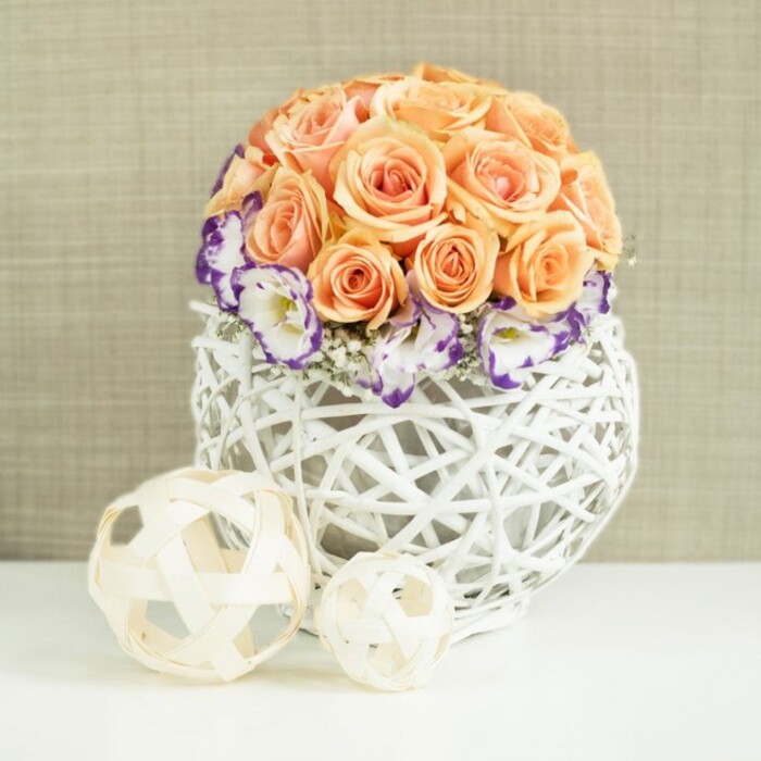 A romantic centerpiece is displayed against a gentle beige fabric backdrop, the kind you might see gracing the head table at a Stanmore wedding or an elegant anniversary dinner at a local restaurant. The arrangement centres on a rounded dome of roses in a spectrum of peach and apricot hues, from blushing champagne to warmed, almost honeyed tones. They are packed closely together so that their velvety petals touch and overlap, forming a soft, cushion-like globe that feels abundant yet refined. Embracing the base of this rose cluster is a ring of lisianthus, each bloom white at its heart but edged with a distinctive, saturated violet-purple rim. These decorative borders give the impression of fine detailing on a luxury fabric, adding depth and a sense of artistry. Between the lisianthus blooms, diaphanous clusters of white gypsophila peek out, their tiny star-shaped flowers offering a light, cloud-like counterpoint to the larger blossoms. All of the flowers are secured in a broad, bulbous container woven from white rattan or wicker strands. The open lattice design allows air and light to pass through, creating a delicate interplay of lines and shadows on the white surface below. It feels handcrafted yet contemporary, a piece that would blend effortlessly into stylish homes along Uxbridge Road or near Stanmore Country Park. In front of this main vase, two decorative orbs made from pale wooden strips are placed on the table, one medium-sized and one smaller, their interlaced patterns mirroring the weave of the container. The entire composition-soft roses, vivid purple edges, airy filler flowers, and natural woven textures-creates a mood of calm sophistication and timeless romance, perfect for capturing the essence of special occasions in Stanmore.