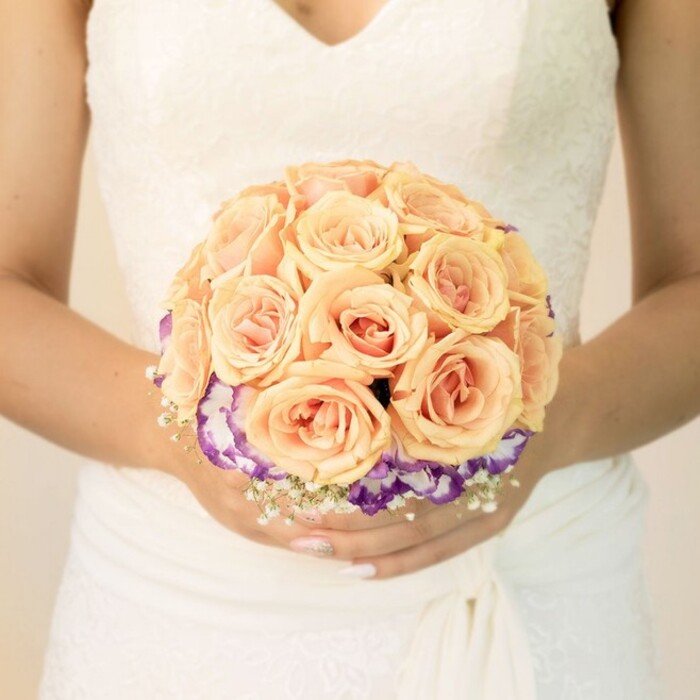 In this detailed, chest-level photograph, the focus rests on a bride's hands as they gently support a round bouquet, her torso framed against a softly blurred, light background that suggests the calm of a Stanmore morning. She wears a traditional white gown with an ivory hint, the bodice decorated with fine lace that traces a modest V-shaped neckline. The gown's fabric gathers smoothly around her waist before cascading in quiet folds, providing a graceful canvas that allows the colours of the flowers to truly stand out. The bouquet itself is meticulously crafted, its upper section dominated by numerous peach-toned roses arranged in a tight, domed cluster. Each rose transitions through a gentle gradient-from pale apricot at the outer petals to deeper, blush-coral tones nested in the center-creating a sense of warmth and romantic depth. Just beneath this rose-filled crown, a ring of ruffled blooms, most likely lisianthus, introduces striking contrast. Their petals are edged in rich, velvety purple that gradually fades to luminous white in the middle, forming a vibrant band of colour around the base of the peach roses. Fine sprays of delicate white filler flowers, reminiscent of baby's breath, peek out around the lower edge of the bouquet, adding a cloud-like softness and classic bridal charm. Her nails are polished in a pale, understated shade with a subtle glimmer on one thumb, a small but personal touch that echoes the bouquet's blend of tradition and individuality. The overall mood is one of poised celebration, the kind of carefully considered floral design you might expect from an experienced local florist preparing arrangements for a Stanmore wedding, whether in a nearby church, a family garden, or a favourite reception venue.