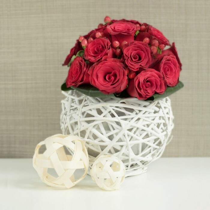 A lush, tightly gathered mound of deep red roses and berries sits atop a pristine white surface, its rounded silhouette creating a classic wedding table profile that feels right at home in Stanmore's elegant venues. The roses are the star: densely packed, their velvety petals varying from dark ruby at the centres to slightly softer crimson at the edges. Some blooms are in the first flush of opening, with neat, compact spirals, while others are more generously unfurled, giving the arrangement a sense of gentle movement. Nestled between the roses are small clusters of red Hypericum berries, their smooth, bead-like forms and high gloss adding a tactile counterpoint to the softness of the petals. A subtle band of dark green foliage hugs the base, glimpsed only in small flashes, yet adding depth and a touch of natural woodland colour beneath the dominant reds. Supporting the bouquet is a unique spherical vase crafted from white-painted twigs or wicker, woven into an open lattice that wraps around the base of the blooms. The white frame provides a crisp contrast to the rich flowers, its airy structure allowing light to filter through and cast delicate shadows on the white tabletop below. To the left of the arrangement, two decorative spheres made from natural, pale wood strips echo both the round shape and the woven texture of the vase, their different sizes introducing a relaxed, harmonious rhythm. The backdrop is a softly blurred wall in warm beige or taupe with a subtle, woven-like texture, ensuring nothing distracts from the floral focal point. The overall effect is intimate, romantic, and quietly luxurious-an ideal table arrangement for weddings, anniversaries, or refined dinner parties in homes and venues throughout Stanmore.