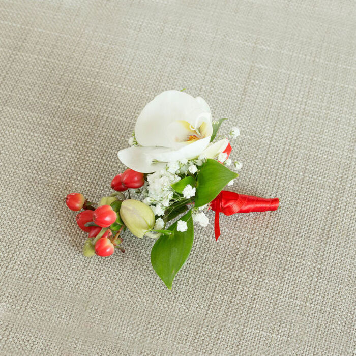 A refined floral buttonhole is shown from a slightly raised, overhead angle, resting on a pale beige, woven textile background that feels like natural linen, its gentle texture adding warmth without drawing attention from the flowers. The central bloom is a pristine white orchid, most likely a Phalaenopsis, its wide, velvety petals opening gracefully to reveal a throat washed with soft creamy yellow and a hint of muted orange, creating a subtle heart of colour within the otherwise ivory flower. Encircling the orchid, clusters of tiny white baby's breath blossoms gather in airy sprays, each little floret contributing to a misty, cloudlike effect that frames the central bloom and adds romantic volume. Nestled alongside these delicate whites is a compact grouping of shiny, round red berries, reminiscent of hypericum, their glossy skin catching the light and offering a jewel-like contrast. Interspersed near the berries are a few smaller, green berry buds that look freshly picked, bringing a sense of new growth and natural variety. Around the flowers and berries, layered green foliage fans out in different shapes: some leaves are broader and more pointed, others slim and elongated, the mixed textures creating movement and depth, similar to the foliage arrangements often seen in window displays along Church Road in Stanmore. The stems of all these elements are gathered tightly and wrapped with a bright red satin ribbon, the fabric wound smoothly and tied into a small, precise knot that gives the design a polished finish, as though prepared moments ago in the workroom of Flowers Stanmore. This carefully crafted boutonniere, with its balance of white orchid elegance, festive berries, and fresh greenery, suggests a special occasion such as a winter wedding, Christmas party, or black-tie event in the Stanmore area, offering a small but striking touch of nature to complement a formal outfit.