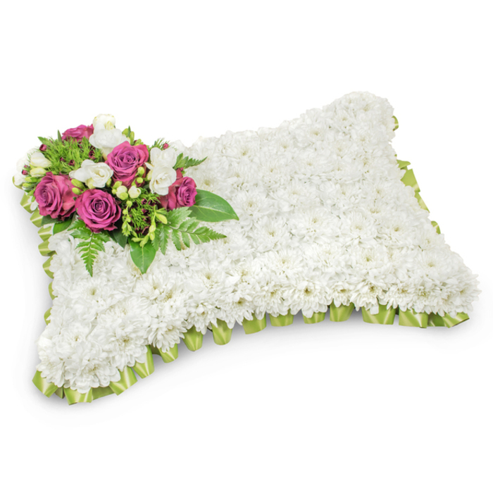 Seen from a gentle angle, this memorial pillow is a study in serene contrasts: a broad, rectangular cushion entirely cloaked in small white chrysanthemums, their compact heads forming a soft, uniform field that suggests purity, comfort, and everlasting sleep. The white surface is deliberately unbroken except for a composed accent in the top-left corner, where several deep fuchsia roses rise like well-tended jewels-rich, saturated centres wrapped in velvety petals. Interlaced among these roses are sprays of white freesias and tiny white buds that add airiness and a whisper of fragrance, their delicate forms lifting the heavier rose tones. Beneath and around the cluster, slender fern fronds and polished leafy greens create a layered understorey, lending sculptural depth and a gentle sense of movement that keeps the eye engaged. The edge of the pillow is finished with a looped, light green satin ribbon, pleated with precision so its muted sheen frames the piece without distraction. Photographed against a stark white backdrop that isolates the composition, this tribute reads as both a crafted keepsake and an emblem of condolence-meant for viewing at a wake, funeral service, or graveside. As a local bouquet maker who has arranged similar cushions for services at the parish on Stanmore Broadway and for ceremonies near Stanmore Tube, I aim for balance, scent, and proportion so the arrangement offers comfort and quiet dignity to those who gather to remember.