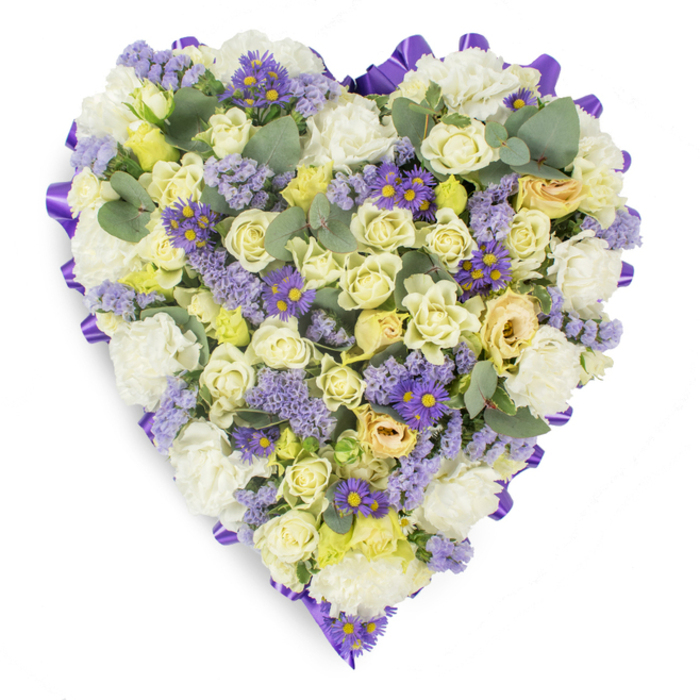 Handcrafted to convey respect and remembrance, this heart-shaped floral tribute is densely assembled on a stark white studio background and neatly outlined with a folded purple satin ribbon that forms a clear, elegant perimeter. The mass of blooms is a soft symphony of white, cream and pale yellow - predominantly cream and pale yellow roses whose tightly furled petals create layers of plush, tactile depth. Interlaced among them are abundant white carnations, their ruffled faces adding weight and a comforting, classic presence to the arrangement. Delicate clusters of lavender statice and small, vivid purple aster-like flowers with golden centers scatter through the flowers like notes of memory, giving the design subtle rhythm and points of visual focus. Rounded, blue-green eucalyptus leaves punctuate the composition, their silvery undersides and distinctive shape providing gentle contrast and a suggestion of fresh, aromatic greenery. The overall finishing is precise yet tender, illuminated by soft studio light that highlights petal contours and the gentle gradations between cream and pale green in some blooms. As a local florist based near Stanmore Broadway and the Jubilee line, I picture this tribute arriving for a funeral or wake in the Stanmore area, its sober palette and structured heart shape speaking clearly to mourners: an emblem of lasting affection and tranquil consolation. The piece feels both timeless and approachable - a considered floral message of love, sympathy and quiet devotion suitable for graveside placement, a chapel display, or as a condolence gift delivered with care.