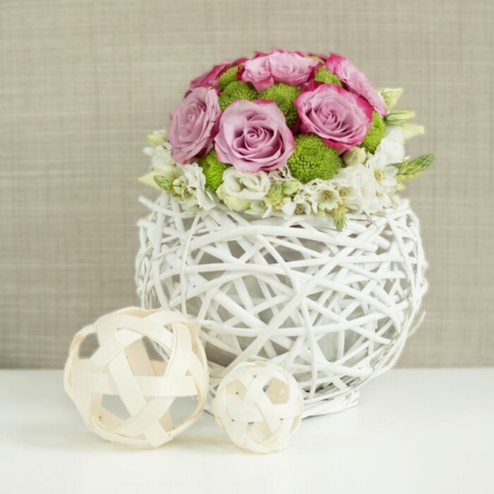 This image presents a graceful floral arrangement that would feel perfectly at home on a wedding table in Stanmore, perhaps set in a light, airy room overlooking tree-lined streets. The arrangement sits on a flawless white tabletop, drawing the eye first to its unusual vase: a round, low sphere woven from slender white branches or twigs. The material twists and overlaps to form a delicate lattice, leaving open spaces that give the piece an airy, sculptural quality. Nestled within is a dome-shaped bouquet of roses in soft pink and lavender hues. Many blooms are fully open, displaying layered petals that curl outward in gentle spirals, while others are slightly less open, adding variety to the texture. Some petals deepen into a richer pink at their edges, providing subtle contrast and visual interest. Among the roses, small green button chrysanthemums are carefully placed, their tightly packed petals and rounded form contributing a lively pop of colour and a pleasing textural counterpoint. Around the lower edge of the dome, dainty white filler flowers-perhaps baby's breath or similar delicate blossoms-emerge between the larger blooms and softly drape toward the rim of the vase, blurring the line between bouquet and container. To the left of the vase, two decorative spheres of pale wicker or rattan rest on the tabletop, one larger, one smaller, both with open, interwoven strands that echo the look of the vase. The background is rendered in soft focus, a warm beige fabric with a subtle texture that recalls linen or burlap and keeps attention settled on the flowers. The overall mood is one of quiet luxury and gentle romance, suited to weddings, anniversaries, or special gatherings in Stanmore homes and venues.