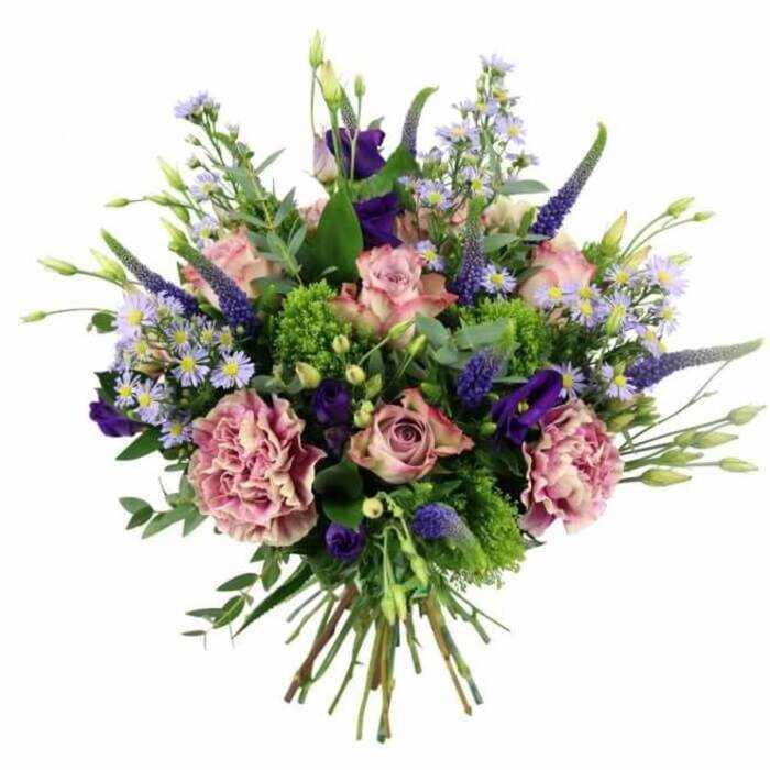 A carefully crafted hand-tied bouquet is displayed against a spotless white background, its colours harmonising in soft pinks, mauves, purples, and greens that feel very much at home in the tranquil surroundings of Stanmore. The bouquet has a classic rounded silhouette, with the blooms arranged so they spill outward in a loose yet deliberate dome. At the centre, several pastel pink roses form a romantic focal point. Each rose displays layered petals, some tightly wound and others fully open, with colour that gently transitions from a deeper blush in the middle to almost ivory along the outer edges. Interwoven with these roses are large carnations in dusty rose and muted mauve, their delicately frilled petals creating a billowy, almost cloud-like texture that softens the entire arrangement. Tall, slender spikes of deep violet-blue Veronica rise subtly through the flowers, providing vertical interest and drawing the eye upward, not unlike the elegant lines of the nearby church spires or mature trees around Stanmore Hill. Scattered generously throughout are small, star-shaped asters in light lavender and periwinkle tones, each brightened by a dainty yellow centre, giving the impression of a soft haze or floral mist. Deep purple lisianthus blooms add further dimension, their slightly ruffled, bell-like forms and tight buds echoing the gentle curves of the roses and carnations while enriching the colour palette. A lush collar of greenery surrounds the flowers, featuring cool, silvery eucalyptus leaves, bright chartreuse-green filler that could be sedum or green trick dianthus, and other assorted foliage that brings structure and freshness. The stems below are cleanly cut and tightly bound, showing the precision of an experienced florist at Flowers Stanmore. The overall feeling is one of thoughtful elegance and quiet joy, ideal for imagining a bouquet delivered to a dinner table near Stanmore Country Park, to a loved one recovering at home, or to celebrate a personal milestone with subtle sophistication.