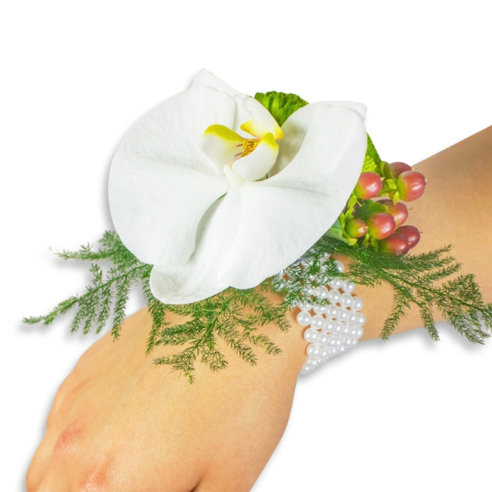 A refined wrist corsage photographed on a person with fair skin, set against a stark white background that emphasizes every curve and texture. At its centre sits a single white Phalaenopsis orchid - broad, unfurling petals smooth as silk, with a bright yellow labellum and a richer orange-yellow throat that forms a small, luminous focal point. Beneath and around that central bloom, airy sprigs of asparagus fern spread in soft, bright-green fans, their fine fronds creating a delicate lacework that supports and lightens the composition. Tucked to the upper right of the orchid, a compact cluster of Hypericum berries provides tonal contrast: glossy, round fruits shifting from light green through rose to a full cranberry hue, conveying a sense of seasonal depth. A sliver of ruffled lime-green accent foliage near the berries introduces additional texture and a contemporary note. The arrangement is mounted on a multi-strand bracelet of tiny white pearls, each bead catching the light and resting elegantly against the wrist - an interplay of tactile coolness and floral warmth. The overall impression is quiet luxury: soft, lightly scented, and artfully balanced for formal occasions such as weddings, proms, or milestone celebrations. As a local Stanmore florist familiar with Harrow Weald and the leafy lanes around Canons Park, I design pieces like this to be comfortable for long wear and flattering in photographs, with a composition that reads equally well in daylight and soft evening light.