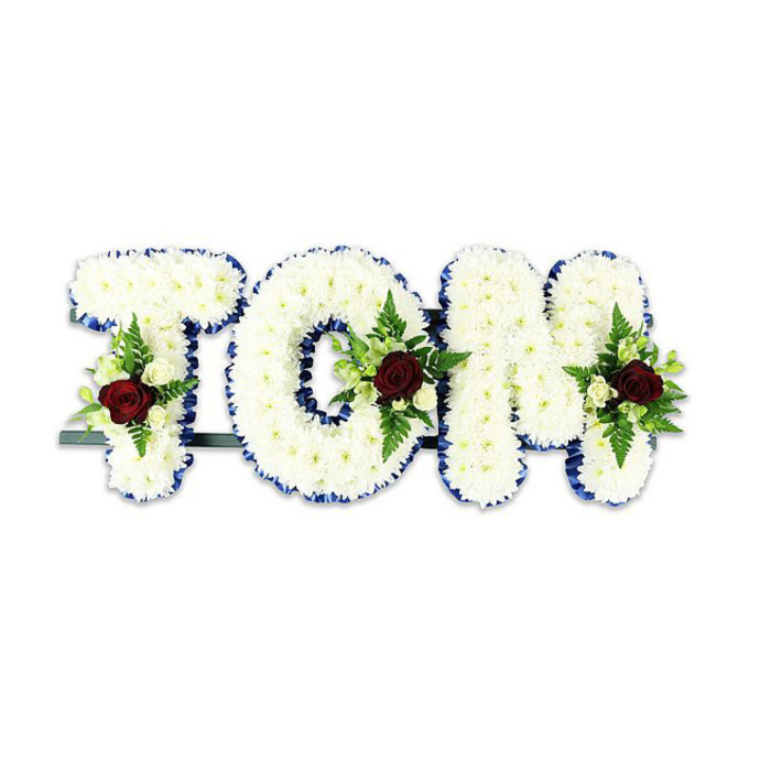Presented against a stark white surface, this bespoke sympathy arrangement spells the name TOM in substantial three-dimensional letters, each letter densely sheathed in small, immaculate white chrysanthemums that form a soft, tactile surface. The precise edges of the letters are outlined with a vivid royal blue ribbon that provides a crisp graphic frame and a stately contrast to the gentle whites. Within each letter, a composed floral spray adds focal sentiment: a single, richly hued dark red rose sits at the heart of every cluster, its velvety petals suggesting deep affection and solemnity. Surrounding the red focal roses are diminutive ivory spray roses and tight buds, which introduce a delicate, sweet scent, and swathes of fern fronds and mixed greenery that bring structure, depth and a rustling, leafy texture. A narrow dark green or black metal rail is discreetly mounted at the rear to support the letters for upright display - a practical detail that ensures the tribute remains steady during a funeral service or wake. The piece reads as both an assertive message and a gentle embrace: the white chrysanthemums speak of purity and comfort, the royal blue edge of the ribbon lends dignity, and the red roses convey enduring love. This design would be suitable for memorials held in local venues across Stanmore and nearby Harrow, crafted to be both visually distinct and profoundly respectful.