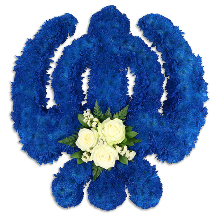 A large Khanda symbol crafted entirely from flowers forms the centre of this composition, representing Sikh faith and identity with unmistakable clarity. The tribute is photographed against a bright white background, which throws the deep royal blue of the chrysanthemums into sharp relief. Each component of the Khanda-the encompassing chakkar, the strong upright double-edged sword, and the two mirrored kirpans-has been painstakingly traced by densely packed, small-headed blooms, giving the surface a tactile, cushion-like quality. The blue tone is intense yet soothing, conveying solemnity without heaviness, in a way that feels at home in the calm atmosphere of Stanmore's places of worship and reflection. Nestled at the base of the central sword is a contrasting detail that softens the design: three pristine white roses, poised at full openness, sit surrounded by tiny white filler flowers and encircled by dark green foliage. This smaller cluster feels intimate within the larger emblem, like a personal message of love and remembrance held within the broader symbolism of Sikhism. Subtle shadows fall beneath the petals and along the floral contours, highlighting the three-dimensional form created by careful hands. Imagined as a bespoke piece from skilled florists serving families across Stanmore, this arrangement would be particularly suited to Sikh funerals, memorials, or anniversary remembrances, whether placed in a gurdwara off London Road or at a crematorium chapel nearby. Its interplay of vibrant blue, snow-white blooms, and lush greenery offers a refined way to honour a loved one's journey, blending cultural significance with the comforting presence of fresh flowers.