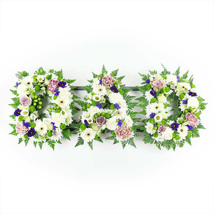 This photograph captures a refined funeral flower tribute spelling out DAD in three capital letters, arranged in an orderly horizontal row and centred against a vivid white backdrop. The background's simplicity allows the floral work to command full attention, highlighting the craftsmanship in every stem. Each letter is composed primarily of snow-white chrysanthemums that are tightly clustered together, creating a plush, uniform base. Their rounded petals overlap to form a soft, inviting texture, while white gerbera daisies punctuate the design, their distinctive dark centres introducing subtle depth. Nestled between the larger blossoms are smaller white flowers-lisianthus, spray roses, and perhaps a few miniature chrysanthemums-introducing fine, lace-like details that make the letters feel rich without becoming fussy. Around the outer contours, an abundance of fresh green foliage provides a natural frame. Wispy fern fronds trace the curves and lines of each letter with gentle precision, while pockets of bright green button chrysanthemums or berry-like accents sit among the leaves, echoing the new growth found in nearby Stanmore Country Park and Bentley Priory Nature Reserve. Scattered throughout the letters, carefully chosen accent blooms in shades of purple break the monochrome scheme. Deep violet flowers, reminiscent of pansies, violas, or spray carnations, add a noble, contemplative tone, while softer lavender and mauve roses and carnations bring warmth and tenderness, hinting at treasured memories and family stories. The overall mood of the arrangement is calm and reverent, a design that would sit beautifully beside a coffin in a Stanmore chapel or at a graveside service. It offers a personalised, floral expression of love and remembrance for a father, created with the quiet attention to detail that local families appreciate.