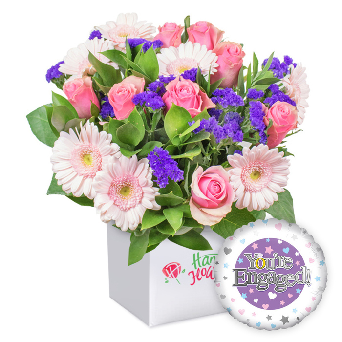 An exuberant pink bouquet rests in a snowy white square box, the clean geometry giving the blooms room to breathe. A cluster of full-bloom pink roses occupies the heart, their petals curling in soft spirals that emit delicate perfume and romance. Nearby, light pink gerberas with white-tipped petals sparkle like little suns, their centers pale yellow and inviting. Against this rosy chorus, clusters of deep purple statice peek out, adding depth and a regal contrast. Framing the stems, variegated green foliage spills softly, creating a lush, garden-like texture that feels fresh and natural. The box's front bears a slender red rose emblem and green script, marking a place of care and local craft. To the lower right, a glossy engagement-themed foil balloon completes the scene. The balloon features a central purple circle with 'You're Engaged!' in bright silver, the 'O' in You're stylized as a gleaming golden engagement ring. The outer rim is white, liberally dotted with tiny pastel hearts and sprinkled with silver stars, echoing the joy of a first big milestone. This arrangement embodies a thoughtful Stanmore-made gift-elegant, romantic, and ready for delivery to a newly engaged couple's home, workplace, or a weekend visit to Stanmore's parks and cafés to celebrate.