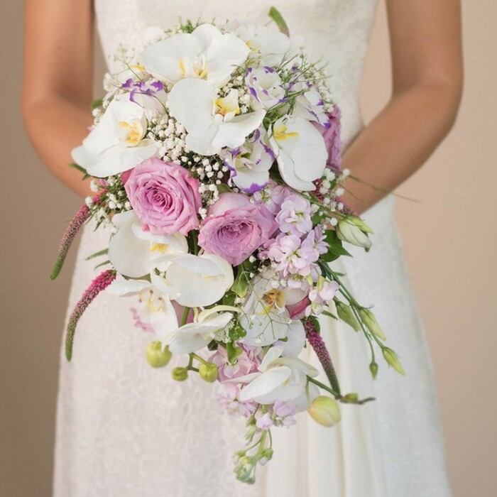 Artfully arranged by Flowers Stanmore, this cascading bridal bouquet represents the spirit of Stanmore weddings-classic, fresh, and touched by local tradition. The image centers on a bride, her upper body enveloped in a white or ivory lace-adorned gown, delicately holding an exquisite arrangement. Majestic white Phalaenopsis orchids with their golden-yellow throats form the heart of the bouquet, framed by generously blooming blush pink and lavender roses, each petal soft and richly textured. Ethereal clusters of white gypsophila thread between the showier blooms, lending a sense of weightlessness, as if a spring breeze from Stanmore Common were stirring through the bouquet. Magenta amaranthus trails enthusiastically, its plush, dramatic tendrils evoking movement and flair. Intermingled are small lavender blossoms, perhaps sweet peas or stocks, adding layers of pastel color and subtle scent. Sprigs of fresh foliage and slender, unopened flower buds emerge among the blooms, imbuing the arrangement with renewal and vibrancy. The bride's hands are placed with gentle intention at her waist, her bouquet forming both the centerpiece of the image and a symbol of hope, devotion, and artistry. Against a soothing neutral background, the rich palette and expert craftsmanship shine-an ideal inspiration for Stanmore couples envisioning elegant, memorable wedding flowers close to home.