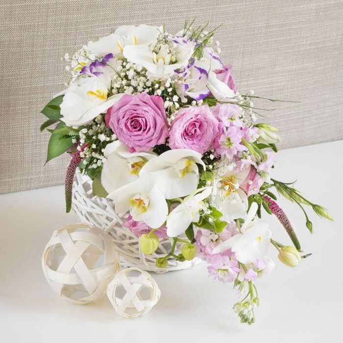 An elegant floral centerpiece of pink and white blossoms is displayed in a round white wicker basket, the kind of design that could easily grace a wedding table or family celebration in Stanmore. The arrangement is lush and full, with soft natural lighting emphasizing the subtle variations in colour and texture. Prominent at the front and centre are pastel pink roses, opened wide so that each petal can be seen curling softly away from the core, creating intricate spiral patterns that suggest romance and care. Interlaced with the roses are luxurious white Phalaenopsis orchids, their broad, smooth petals arching gracefully outward. Each orchid reveals a delicately patterned center in tones of yellow and warm apricot, dusted with a hint of mauve, giving the overall bouquet a rich, high-end bridal feel. White Lisianthus blooms provide an additional layer of refinement, their slightly ruffled petals-some edged with a gentle lavender hue-offering a soft, fluttering contrast to the bolder rose and orchid shapes. Tiny white baby's breath flowers are woven throughout, scattering points of delicate light and giving the arrangement a soft, dreamy halo that would feel at home in a reception space near Stanmore Broadway. Pale pink stock flowers, clustered along upright stems, peek above and spill just beyond the basket rim, adding vertical interest and a sense of gentle movement. Fresh greenery, including slim leaves and emerging buds, punctuates the design and creates a lively balance with the pastel blossoms. On the pristine white surface beside the basket, two decorative woven spheres crafted from pale natural material rest in quiet symmetry, their intertwined strands echoing the rustic charm of the basket. In the background, a softly textured light beige or taupe fabric creates a warm, neutral canvas that keeps the focus squarely on the flowers. The overall impression is of thoughtful craftsmanship, calm celebration, and understated luxury, ideal for marking special occasions throughout Stanmore.