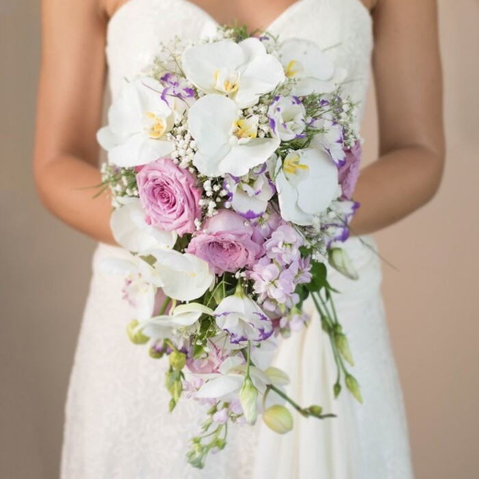 Framed elegantly in this vertical close-up, a Stanmore bride gracefully holds a sumptuous cascading wedding bouquet-a refined floral piece grounded in local artistry and natural beauty. The muted, softly blurred beige background underscores the intricate detail of the arrangement: prominent snow-white phalaenopsis orchids stretch their wide petals, each with subtly glowing yellow throats, commanding center stage. Interwoven among them, textured blush-pink garden roses, their ruffled forms lush with layers, evoke romance and gentle nostalgia, while smaller lavender and light purple lisianthus create pockets of depth and sophistication. Fragrant sprigs of purple stock and miniature white gypsophila (baby's breath) are thoughtfully dispersed, lending airiness and bold dimension, complemented by slender threads of green foliage. The bouquet's artful teardrop silhouette gently cascades downwards, echoing the lines of chic bridal fashion seen in Stanmore's elegant venues. Behind the florals, a glimpse of a fine strapless lace gown reminds one of family traditions and timeless ceremonies held near Stanmore Common. The overall effect is one of luxury and serene grace-a captivating wedding bouquet meticulously curated for North London brides by local experts, perfect for those marking life's most precious milestones in the Stanmore area.