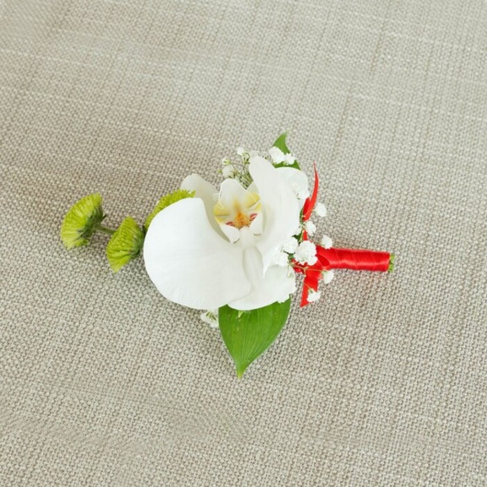 Set against a softly woven, light beige linen background, this sophisticated boutonniere appears almost like a small work of art carefully laid out on a dressing table before a ceremony in Stanmore. The fabric beneath has a natural, slightly textured weave that adds warmth without distracting from the floral design. At the centre, a single white phalaenopsis orchid blooms in full, its broad, gently arched petals forming a flawless, sculptural frame. The petals are creamy white with the faintest suggestion of shadow at their edges, while the orchid's throat glows with soft yellow, merging into fine reddish-orange speckles and lines that lend an intricate, painterly quality. Surrounding the orchid, clusters of delicate white baby's breath give the piece an ethereal halo, their tiny blooms reminiscent of delicate lace or drifting blossom in springtime around the quiet streets near Stanmore Hill. To the left, two compact lime-green chrysanthemum buds, spherical and covered in small, pointed petals, provide a vivid counterpoint, adding freshness and a contemporary twist to the otherwise classic palette. A rich, deep green leaf cradles the entire arrangement from below, visually grounding it, while a smaller, darker leaf tucked near the base brings subtle depth to the foliage. The stems are gathered and wrapped with meticulous care in a bright crimson red satin ribbon that spirals down into a neat, understated bow, its glossy surface catching the light and injecting a celebratory note. The overall effect is timeless and refined-a boutonniere perfectly suited to a groom or groomsman arriving at a local church or venue, perhaps selected from the curated wedding designs offered by Flowers Stanmore for same-day events across the HA7 area.