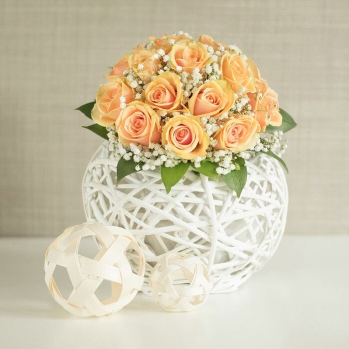 This image presents a refined, contemporary floral arrangement that would feel perfectly at home in a Stanmore living room overlooking the greenery near Canons Park. At the heart of the composition is a domed bouquet of roughly two dozen peach and apricot roses, their blossoms gathered so closely that the petals almost seem to lean on one another. The inner petals glow with a deeper, warm orange, gently transitioning to softer, more pastel tones at the edges, giving the bouquet a sun-kissed look, as if caught in gentle late-morning light. Interlaced throughout are numerous airy tufts of white baby's breath, each tiny flower a pinpoint of brightness that softens the density of the roses and adds a lovely, romantic haze. Deep green leaves, discreetly tucked beneath and between the blooms, introduce a natural depth and prevent the arrangement from feeling too sweet or monochrome. The flowers are cradled in a large spherical vase fashioned from white woven strips in an open lattice design; the crisscrossing framework feels both sculptural and delicate, allowing glimpses of stems while keeping the overall effect clean and modern. In the foreground, on a pristine white tabletop, two smaller decorative orbs rest side by side, crafted from pale, natural-looking material and echoing that same woven texture, subtly reinforcing the design theme without overshadowing the flowers. The background is a soft, light beige or greige wall with the faintest hint of vertical texture, which gently diffuses the light and keeps the focus on the bouquet. Everything about the scene speaks of calm elegance and thoughtful styling, the sort of centrepiece one might choose for a small wedding, an anniversary dinner at home, or stylish home staging in and around Stanmore.