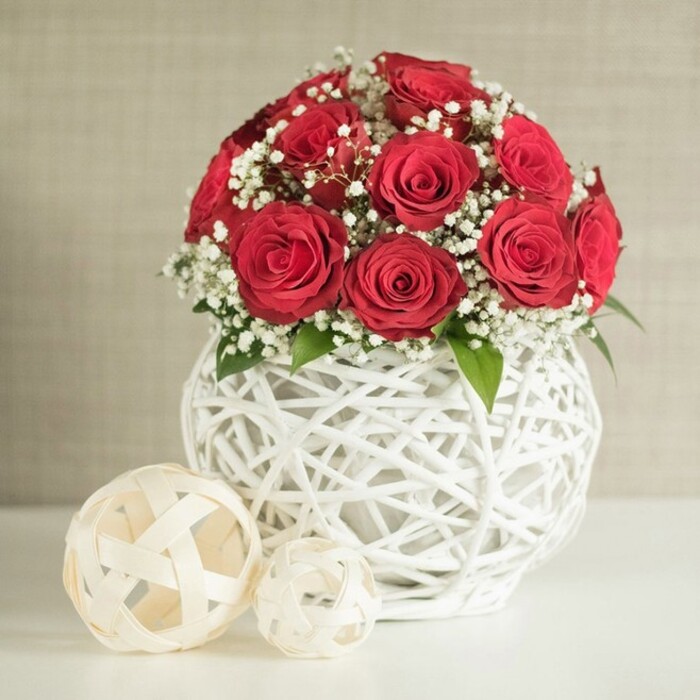 In this elegant still life, a voluminous bouquet of red roses forms a striking central dome, the blossoms nestled closely together so their velvety petals overlap in a rich expanse of crimson. Each rose is fully opened, its petals curling outwards in precise, layered spirals that showcase the flower at its most opulent stage. Between these passionate red blooms, clusters of white baby's breath weave gently through the arrangement, their minuscule flowers scattering points of brightness across the darker tones and creating a soft, airy counterpoint. Subtle touches of green foliage emerge here and there, providing natural breaks in the colour and lending the composition a sense of freshness and life. The bouquet is supported by a distinctive, bulbous vase crafted from white strands that wind around each other in an open, interlaced pattern. This airy construction, reminiscent of white rattan or contemporary wickerwork, gives the container an artistic, handcrafted quality while allowing the background light to filter through. The vase rests on a smooth, white tabletop, its simplicity emphasizing the textures above. To the left of the main arrangement sit two woven decorative spheres in a light cream hue, one notably larger than the other, each constructed from flat strips that crisscross in an open lattice, echoing the form and feel of the vase. Behind, a neutral beige wall with a soft, fabric-like texture fades gently out of focus, keeping the viewer's attention on the flowers and their sculptural container. The image evokes a carefully planned table setting in a Stanmore home or event space near Canons Drive, well suited for wedding receptions, Valentine's dinners, or thoughtful anniversary surprises delivered locally.