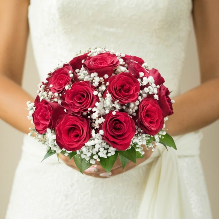 This evocative portrait, set amid the tranquillity of Stanmore, showcases a bride's hands cradling an artfully composed bouquet-a true celebration of time-honoured romance. Centred in the frame is a dome of deep red roses, their petals rich, velvety, and perfectly shaped. Interlacing these dramatic blooms are abundant clusters of white gypsophila, or baby's breath, each minute blossom contributing an ethereal softness that offsets the intensity of the reds. Emerald-green leaves emerge from the bouquet's base, their freshness reminiscent of Stanmore's green spaces and gardens. The bride's hands, with their delicate manicure, enfold the stems with quiet assurance, while the white or ivory wedding dress behind them glimmers with lace detail or fine embroidery. A gentle drape or sash falls from one shoulder, creating a sense of movement and elegance that pays homage to bridal tradition. All around, the background melts into a creamy blur, ensuring nothing distracts from the floral artistry and anticipation of the moment. Ideal for those dreaming of elegant weddings in Stanmore, this bouquet is a testament to local craftsmanship and timeless love.