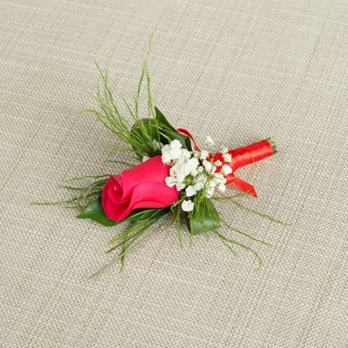 Set against a softly textured, natural beige fabric background that resembles fine linen or rustic burlap, this beautifully composed boutonniere appears as if laid out on a dressing table before a special event in Stanmore. The arrangement is angled gracefully, pointing towards the upper right, giving the piece a dynamic yet poised presence. At the heart of the design is a single rosebud in a striking fuchsia or deep magenta pink, its petals tightly wrapped around one another in a perfect spiral. The rose's surface is velvety and smooth, with subtle shading in the petals that suggests depth and freshness, as though it has just been selected from a florist's bucket that morning. Surrounding the rose are delicate clusters of white baby's breath, each cluster made up of tiny, rounded blossoms that form a frothy halo, softening the boldness of the pink and adding a romantic, cloud-like texture. Supporting this central cluster is a balanced mix of greenery: fine, bright green, feathery foliage radiates outwards, giving the boutonniere an airy, garden-inspired feel, while slightly broader, more solid dark green leaves provide a structured base and visual anchor. At the lower end, the stems are cleanly trimmed and bound together with a vibrant red satin ribbon, meticulously wrapped so that it sits snugly around the stems, with a small, tasteful knot or bow adding a finishing flourish. The ribbon gleams gently in the light, echoing the richness of the rosebud above. This refined, handcrafted lapel flower feels perfectly suited to a groom or groomsman stepping out onto Stanmore Hill or arriving at a local venue, capturing the timeless elegance and personal touch associated with bespoke wedding florals created for North London celebrations.