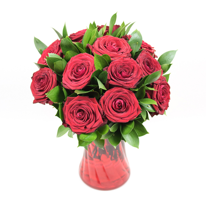 A richly detailed image shows a classic, dome-shaped bouquet of around a dozen to fifteen deep crimson roses arranged in a translucent red glass vase, placed before a spotless white backdrop that heightens every colour and contour. The roses are at that perfect, cinematic stage of bloom: fully open enough to show their spiralling hearts and layered, velvety petals, yet still so full and firm they promise several days of beauty. Each petal folds gently over the next, creating a plush, almost cushion-like texture that makes the red appear even more intense, a true symbol of romance and devotion. Tucked generously among the rose heads, dark, glossy green foliage-likely ruscus or similar hardy greenery-adds body and structure, the elongated leaves emerging between blossoms to break up the red with pockets of cool, natural green. This foliage enhances the bouquet's rounded silhouette, making it feel substantial and thoughtfully balanced rather than crowded. The vase that anchors the arrangement is crafted from clear red glass with a soft, fluted opening and a curving body that narrows gracefully towards the base. Its subtly ribbed or swirled surface catches the light in ripples, so the glass glows with layered tones of cherry and wine, complementing the roses above. The stems inside are faintly visible, contributing to a sense of authenticity and hand-crafted care. Bright, even lighting ensures no detail is lost, from the sheen on each leaf to the shadowed folds of the petals. This bouquet could easily be imagined on a polished hall table in a Stanmore home near Bentley Priory, delivered by a local florist as an anniversary surprise, a Valentine's gift, or a heartfelt declaration of love in the heart of the community.