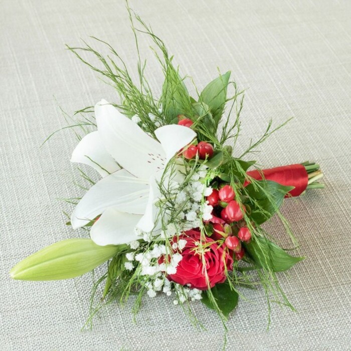 Arranged on a subtle, light beige fabric reminiscent of the refined decor found in Stanmore's peaceful homes, this small bouquet is a tribute to elegance and community values. Dominating the centre, a resplendent white lily flowers proudly, its petals snowy and wide, accented by tiny reddish flecks and golden-tipped anthers which gleam in the soft light. Nestled nearby, a lush, crimson red rose blooms in sculpted perfection, signifying beauty and warmth that resonates with the classic style of Stanmore's leafy avenues. The bouquet is animated by glistening clusters of red berries, their vibrancy effortlessly echoing the festive spirit of the local community. Baby's breath weaves a gentle mist throughout, adding airy charm, while a solitary green lily bud suggests anticipation and promise. Fine strands of green-from feathery ferns to wide, dark leaves-frame the flowers, enhancing the sense of depth and life. The stems are gathered and finished with a sleek, red satin ribbon, bringing unity and polish to the piece. Envisioned for celebrations, especially weddings and anniversaries across the Stanmore area, this bouquet captures both floral artistry and local sentiment in every detail, illuminated by the kind of even, natural light that flatters both flower and memory.