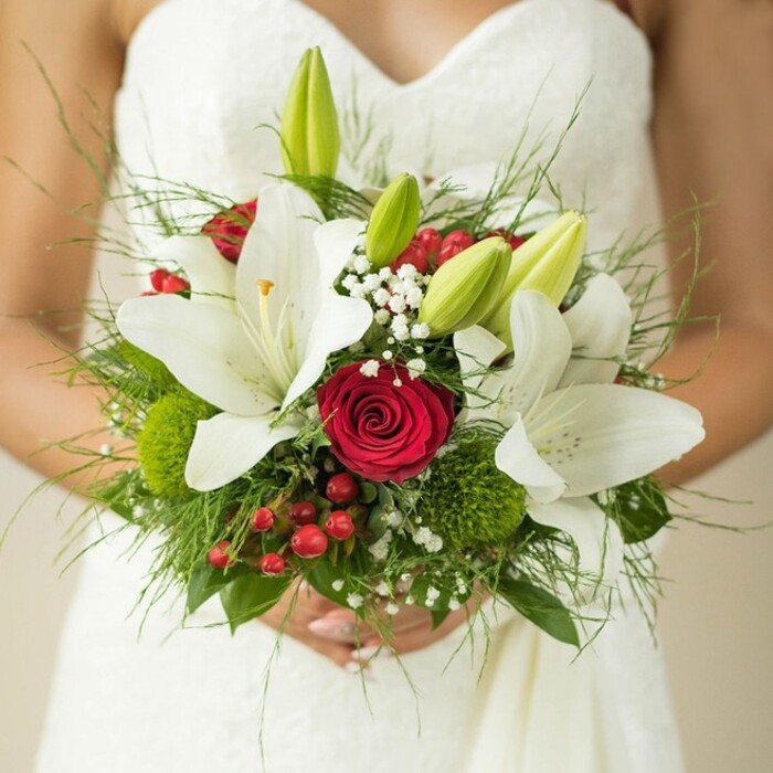 A tender moment unfolds as a bride in Stanmore gently holds an exquisitely arranged bouquet against the soft white folds of her elegant wedding dress. The upper part of her strapless neckline is revealed in the close-up, providing a backdrop for a symphony of blooms: brilliant white lilies with open petals sparkling in the light, cradling golden stamens, and youthful green buds hinting at new beginnings. At the heart of the bouquet sits a single, rich red rose-its tightly packed petals whispering of devotion and romance. Surrounding this focal point are delicate clouds of white baby's breath and dynamic accents from vivid red hypericum berries, adding colour and celebration to the arrangement. Petite green button flowers and a variety of fresh foliage, including feathery ferns reminiscent of Stanmore's local green spaces, imbue the bouquet with dimension and natural beauty. The overall composition suggests both classic elegance and the vibrant, creative flair of the area's florists, making it the perfect inspiration for brides seeking wedding floral designs that reflect Stanmore's unique character and community spirit.