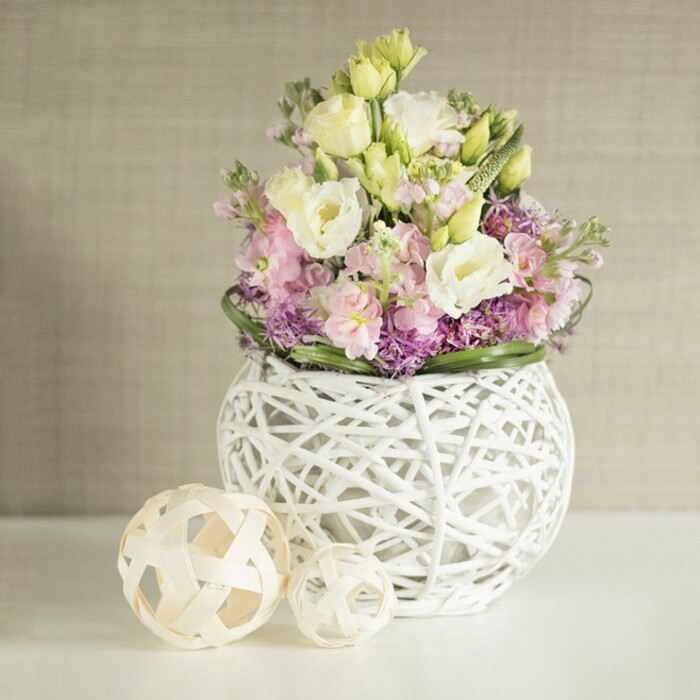 A radiant spring arrangement graces a contemporary white surface in Stanmore, perfectly poised to capture the gentle beauty of North West London's changing seasons. Centered within the composition is a bouquet of creamy white and buttery yellow eustoma, their ruffled petals arranged in soft clusters that evoke the calm mornings in Stanmore's leafy parks. Upward-reaching sprigs of pink stock infuse the bouquet with movement and a touch of chic romance, while below, masses of rich purple minor blooms-likely local asters or alliums-create a lush, color-rich groundwork. A border of deep, fresh green foliage encircles the florals, giving definition and lively contrast, much like the ornamental hedges found around Stanmore Hill. The arrangement is contained within a unique, orb-shaped white vase, its open-weave rattan structure both modern and light, allowing glimpses of the stems and amplifying the freshness within. To its left, two meticulously crafted spheres of pale rattan rest on the table, their open basket designs amusingly echoing the main vessel while adding tactile appeal and artisanal harmony. The backdrop is a soft, textured beige wall-subtle and elegant-ensuring the bouquet's gentle pastels are beautifully highlighted. Infused with the spirit and skill of Stanmore's trusted local florists, this display is a natural fit for wedding tables, anniversary dinners, or anyone wishing to bring a breath of Stanmore's tranquil charm indoors.
