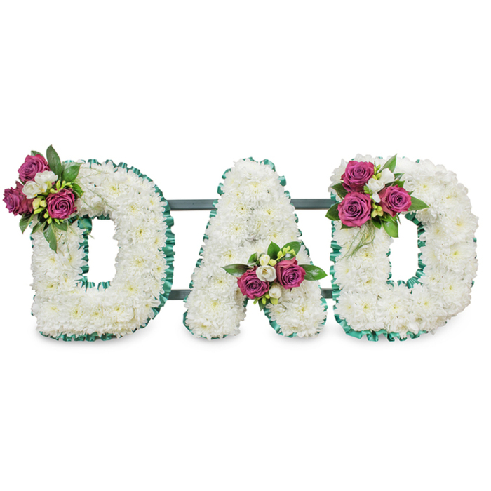 A carefully constructed memorial tribute spells DAD in substantial, three-dimensional letters made from a dense blanket of pristine white chrysanthemums, each tiny floret packed closely to form a seamless, cushion-like surface. Every letter is precisely finished with a vibrant emerald-green pleated ribbon that outlines the shapes and adds a tailored, respectful edge against the white blooms. A discreet dark green support bar runs horizontally behind the letters, indicating a secure, self-supporting design intended for viewing at a chapel, church hall or graveside. Decorative clusters provide the personal accent: the first D is graced with a grouping of deep magenta roses and small white freesia tucked into its upper left curve; the A holds a matching cluster in the lower central V; the final D mirrors the first with a spray on its upper right curve. The roses vary from tight buds to gently opening blooms, their spice-warm scent and velvety texture offering a tender counterpoint to the cool, serene chrysanthemum backdrop. Glossy dark green foliage and tiny white filler blooms sit behind the roses, giving depth and a natural silhouette. Presented against a clean white background in soft, natural-like light, the whole arrangement reads as dignified, intimate and handcrafted by a local shop - Flowers Stanmore - experienced in creating personalised bereavement pieces for services around Stanmore Tube, Stanmore High Street and nearby Canons Park. It speaks of quiet respect, familial love and a thoughtful farewell.