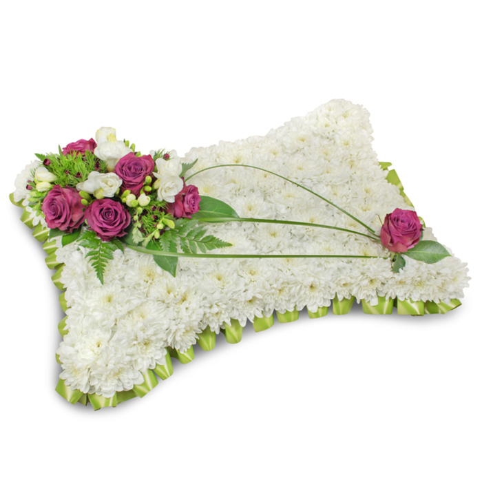 A meticulously fashioned pillow of white chrysanthemums forms the base of this traditional funeral tribute, each tiny floret placed to create a continuous, cloud-like surface that suggests calm and purity. Along the lower edge a neat pleated border of light lime-green satin ribbon runs in a scalloped pattern, giving the cushion a refined frame that lifts the composition with a touch of colour. Dominating the top-left corner is a rich cluster of deep fuchsia roses, their velvety petals unfurling with measured grace, interspersed with delicate white freesia blooms that add a whisper of fragrance and fragile brightness. A generous backdrop of emerald foliage-including feathery fern fronds and broader green leaves-sets the accent flowers off with striking natural depth. Three slender, dark bear-grass blades arch gracefully across the white chrysanthemum field, their sinuous lines drawing the gaze towards the opposite corner where a single, perfectly formed fuchsia rose nests among its own dark leaves, completing the visual balance. The whole piece is photographed against pure white with soft lighting that highlights the textures-the velour of rose petals, the puckered satin edge, the feathery chrysanthemum faces-and the shadow beneath gives it weight and presence. As a seasoned local florist in Stanmore, Flowers Stanmore often styles tributes like this for services at nearby churches or memorials at Stanmore Common, where families seek a composed, respectful expression of condolence that speaks to eternal rest and heartfelt remembrance.