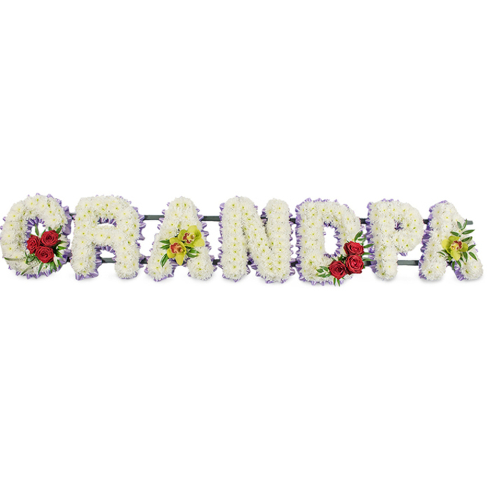 This photograph captures a horizontal floral arrangement that spells out GRANDPA in large capital letters, designed as a funeral or memorial tribute. The word rests on a stark, bright white background, allowing every element of the design to be seen without distraction, as though it were freshly prepared in a quiet Stanmore workshop before delivery. Each letter is built from tightly grouped white chrysanthemums, their small, cushiony flowers layered closely together for full coverage. The result is a consistent, soft texture that looks almost like a padded quilt of petals, conveying warmth and tenderness even in a time of loss. A fine edging of lavender-purple ribbon traces around the outer edges of each letter, providing definition and a delicate hint of colour that gently separates the white letters from their background. Within this field of white, carefully positioned bursts of colour bring additional meaning. Cradled in the curve of the G is a cluster of deep red roses surrounded by lush green foliage, their rich tones suggesting deep love and respect. The first A is adorned with a small spray of yellow-green orchids, the blooms slightly exotic, with elongated petals and subtle markings that speak of individuality and cherished memories. The curved inner space of the D holds another group of red roses, mirroring the G, while the final A carries a second spray of orchids, balancing the arrangement visually. Thin metal rods and a discreet frame support and connect the letters from behind, ensuring the word can be displayed as a single, cohesive piece on an easel or stand at a church in Stanmore, along Marsh Lane, or at a local cemetery. The overall mood is one of calm reverence and affection, honouring a beloved grandfather's life with a clear, heartfelt floral message.