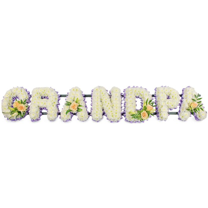 This image shows a carefully crafted floral letter arrangement spelling GRANDPA, designed as a horizontal tribute piece often placed at the front of a funeral or memorial service in and around Stanmore. The seven bold, capital letters stand side by side on a narrow, dark base frame, forming a single, continuous word. Each letter is completely covered with tightly set white chrysanthemums or similar small, rounded blooms. The petals are layered densely, giving the entire surface a soft, cushioned look, as if the letters have been wrapped in a blanket of flowers. Subtle tones of cream or pale yellow at the flower centres hint at warmth beneath the cool white. Encircling the edges of every letter is a refined border of pale lavender or soft lilac blooms, delicately tracing the contours of the G, R, A, N, D, P and A. This pastel outline creates a gentle shadow effect, separating each letter from the clean white backdrop and making the tribute easy to read from a distance. Within the letters themselves, tasteful floral clusters bring additional texture and colour. The G and D are adorned with groups of peach roses and buds, nestled among glossy green foliage and the occasional slender white lily bud. The first and final A each feature a single peach rose framed by fresh greenery, echoing the larger clusters yet keeping the overall balance. The combination of cool white, tranquil lilac, and warm peach suggests peace, fond memories, and enduring familial love. It is the kind of bespoke funeral flower tribute chosen by many Stanmore families to honour a cherished grandfather with quiet beauty and heartfelt respect.