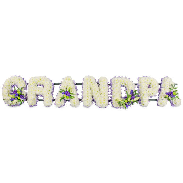In this softly lit image, the name GRANDPA is spelled out in striking floral letters, all in upper case, resting in a neat horizontal row against a simple white background. Each letter is shaped on a sturdy frame and then densely adorned with clusters of white chrysanthemums or carnations, arranged so closely that they form a thick, velvety blanket of petals. The texture is rich yet gentle, evoking a sense of stillness and purity that suits a farewell ceremony held in Stanmore or the surrounding North London suburbs. The unbroken field of white is then thoughtfully embellished with small gatherings of purple and lavender flowers, likely lisianthus or similar blooms, paired with glossy green foliage. These accent flowers are positioned with care: a spray nestling at the base of the G, another at the centre of the first A, more colour placed within the curve of the D and the bowl of the P, and a final cluster softening the last A. The purple tones bring a subtle, regal note to the arrangement, while the greens add freshness and the suggestion of life continuing beyond loss. A narrow dark green support bar runs unseen beneath the letters, quietly connecting them and ensuring the word remains balanced and secure whether displayed atop a coffin, on a stand beside the lectern, or near the entrance to a funeral venue in Stanmore. The tribute has the air of something lovingly planned and carefully executed, perhaps after a family shared memories of their grandfather's kindness and character. It feels like the work of a compassionate Stanmore florist at Flowers Stanmore, translating those stories into blooms that speak of love, gratitude, and a final, gentle goodbye.