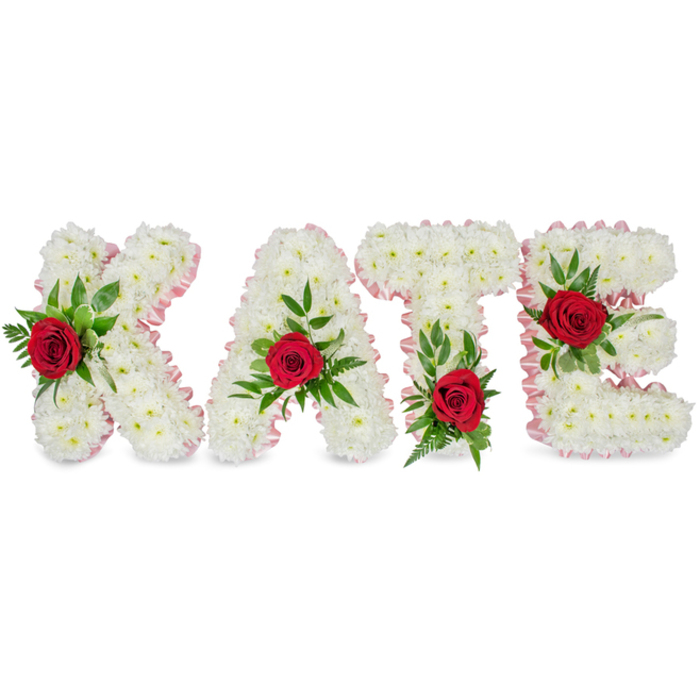 This photograph shows a lovingly arranged floral name tribute spelling KATE in chunky block letters, each letter sculpted from an abundant mass of small, pristine white chrysanthemums whose layered petals form a soft, tactile surface. A delicate pale pink satin ribbon trims the outer edges of each letter, catching light with a faint silk-like glimmer and lending a quiet elegance to the composition. At the heart of each letter sits a single, vivid red rose-deep crimson and velvety-carefully set amid a generous framework of lush green foliage, including broad leaves and feathery fern sprigs that cradle each bloom. The placement is deliberate: the 'K' displays its rose and greenery near the lower part of the left vertical stroke, the 'A' holds its rose at the upper triangular apex, the 'T' is accented by a rose at the base of the left side of its crossbar, and the 'E' features its rose on the middle horizontal bar. Against the clean white backdrop the contrast of colours is striking yet respectful-the white suggesting purity and repose, the red a symbol of love and remembrance, and the green a sign of renewal. The piece radiates a subtle floral perfume and a sense of careful, sympathetic craftsmanship appropriate for a funeral, wake, or commemorative gathering. Locally, such bespoke lettering is often chosen by families around Stanmore Common and the surrounding residential streets when they seek a personalised, dignified tribute that reads clearly from a distance at services and graveside ceremonies.