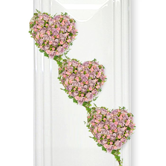 The photograph portrays a delicately crafted sympathy arrangement of three heart-shaped clusters of roses, created with the kind of care and artistry that Flowers Stanmore is known for in the local area. The hearts descend in a gentle diagonal from the top left corner of the frame to the bottom right, forming a graceful, flowing line much like the slow walk of mourners leaving a chapel on Stanmore Hill. Each heart is fully covered in small to medium roses, their petals forming dense layers of dusty lavender and soft blush pink. The colours are muted yet romantic, evoking a sense of quiet tenderness rather than bright celebration. Some blooms appear newly opened, with tight whorls at their centre, while others are more fully unfurled, adding texture and depth that draw the eye inward. Encircling each heart and linking them together is a network of fine ivy strands, their slim green stems looping around the edges and trailing downwards in a natural rhythm. The ivy leaves are glossy and fresh, introducing a vibrant green counterpoint to the pastel flowers and suggesting ongoing life and growth. The entire arrangement stands out against a luminous white background, where faint vertical reflections on the left hint at a polished wall or glass surface, similar to those found in modern funeral venues around Stanmore and nearby Edgware. The light is soft and even, giving the tribute a serene, almost ethereal quality. This triple-heart design feels especially appropriate when a family wishes to symbolise three connected souls or three branches of the same lineage, offering a visual expression of enduring love, remembrance and unity in times of loss.