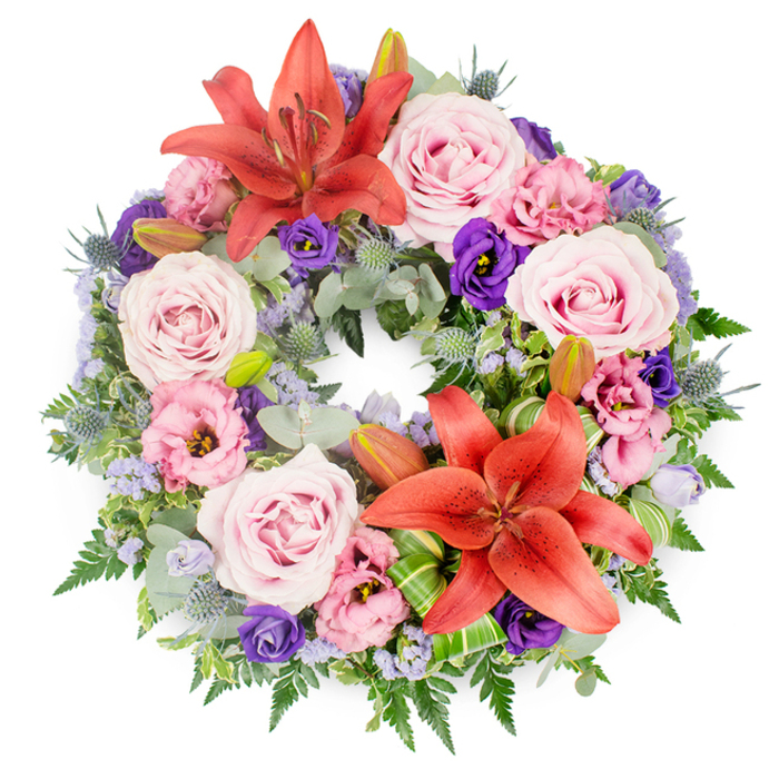 This meticulously crafted circular memorial wreath is shown against a bright white background, every detail clearly visible as if laid out on a table in a Stanmore flower studio. The design is rich but carefully controlled, with colours that move harmoniously from warm to cool. Two large coral-orange Asiatic lilies serve as the design's anchors, one opening fully at the upper centre of the circle, the other placed toward the lower right. Their petals have a subtle sheen and curl gently at the tips, while their stamens add a soft, natural shadow. Between and around these standout blooms, layers of roses in soft blush and lavender-pink form the main body of the wreath. Each rose is fully opened, its spiralling centre giving a sense of depth, like a series of gentle, unfolding memories. Interspersed with the roses are lisianthus flowers in two main tones: a rich, velvety purple that adds gravitas and a lighter, lilac-pink that softens the overall palette. Fine sprays of lavender-blue filler flowers, likely statice or limonium, are worked into the spaces between blooms, creating a delicate haze that ties the colours together. The base of the wreath is a generous collar of mixed greenery. Silvery eucalyptus leaves create a cool, calming foundation, while soft fern fronds and other textured foliage bring a natural, garden-like feel. Blue-grey eryngium thistles, with their spiky, cone-shaped heads, punctuate the greens and echo the cooler tones of the filler flowers. At several points, unopened lily buds rest quietly among the foliage, suggesting life continuing beyond the present moment. Carefully folded bright green leaves near the lower lily display a touch of modern floral artistry. This wreath would sit beautifully at a funeral or memorial service in Stanmore, perhaps adorning the front of a chapel near Stanmore Station or accompanying a hearse through local streets, expressing quiet dignity and heartfelt condolence.