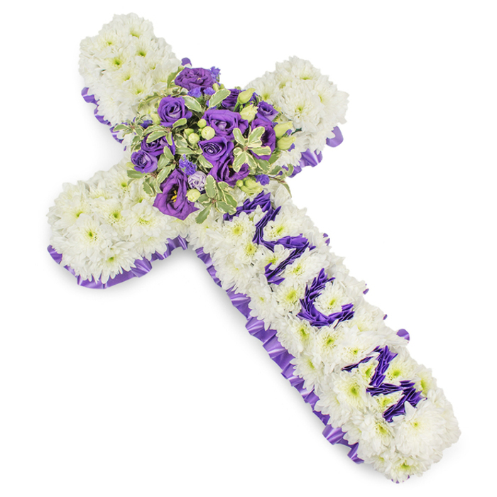 A formally composed Christian cross, photographed on a white backdrop to emphasise every nuanced detail: the body is densely covered in immaculate white chrysanthemum blooms, each floret placed to create a uniform, pillowy texture that suggests calm and purity. The entire perimeter is traced with a rich purple satin ribbon, its ruffle and sheen providing a stately frame that deepens the arrangement's solemn tone. On the upright arm, the word MUM is spelled out in clear, stylised letters fashioned from small purple flower heads-likely eustoma or compact spray chrysanthemums-set to stand out decisively against the white ground. At the cruciform centre a generous focal cluster draws the eye: deep violet roses with tightly furled petals mingle with pale lavender lisianthus, their ruffled edges softening the composition; variegated foliage with lighter margins and darker centres adds layered texture and a hint of spring green, while tiny green berries provide subtle punctuation. The bouquet's cool purples and clean whites suggest reverence and remembrance, the tactile density of blooms offering a comforting solidity. This tribute is designed for use at funerals, wakes, or graveside services in the Stanmore area-suitable for placement by a chapel entrance on Stanmore Hill or beside a hearse on Stanmore Broadway-and speaks of enduring love, respect, and peaceful farewell with restrained, expert floral craft.