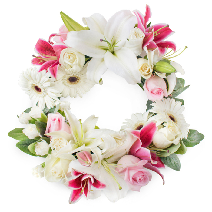 Against a clean white backdrop, a beautifully composed circular wreath of funeral flowers takes centre stage, its open centre surrounded by a profusion of blooms in white, blush, and deep magenta hues. The wreath feels full and thoughtfully structured, with large white lilies providing the strongest visual anchor, their broad, upward-curving petals exposing pale green pistils and golden stamens that add a subtle glow and a sense of sacred calm. Woven among them are vivid deep pink and fuchsia lilies, their star-shaped forms and gentle speckling lending a more dramatic, emotive quality to the composition. Soft pink roses are scattered generously throughout, many in partial bloom or bud, their tightly layered petals hinting at tender memories and unspoken goodbyes. Creamy white roses, fully open, bring classic beauty and amplify the wreath's rounded shape and generous volume. Cheerful white gerbera daisies punctuate the arrangement, their neatly layered petals and dark central discs adding a fresh, modern contrast to the more traditional blooms. Smaller white filler flowers-likely miniature carnations or spray chrysanthemums-fill in any spaces between larger flowers, ensuring the circle appears lush and complete from every angle. Surrounding the floral ring, rich green foliage and leafy sprigs form a soft, natural frame, enhancing depth and the symbolism of renewal. The unbroken circular shape speaks of eternal love and remembrance, making this wreath an appropriate tribute for a funeral or memorial service in Stanmore, perhaps resting near Bentley Priory, a local cemetery, or a quiet family home, conveying heartfelt condolences in a refined and respectful floral design crafted by local hands.