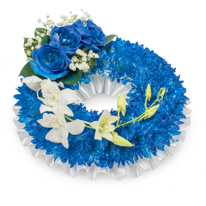 An elegant memorial wreath in deep blues and pristine white, shown against a spare white background so every botanical detail reads clearly. The wreath's body is tightly covered with countless small, royal-blue chrysanthemum-like flowers, their clustered petals producing a lush, textured surface that feels almost cushiony to the eye. A neatly pleated white satin ribbon forms the inner border, its soft sheen and ruffles providing a refined contrast to the saturated blue mass. Near the upper-left curve sits a compact cluster of three striking blue roses, fully open and velvety to the sight; their sapphire petals are complemented by tiny clouds of white baby's breath and a few darker green leaves, creating classical accents that steady the composition. From the mid-left the eye follows a gentle sweep of immaculate white orchids - with the clean lines and rounded lips of Phalaenopsis-style blooms - which cascade toward the lower right and add an airy, graceful motion. Among the orchids, pale green buds at various stages of opening introduce a sense of life continuing even in sorrow. The overall mood is serene and respectful: the deep blues suggest thoughtful remembrance, while the pure whites convey sympathy and peace. This is the kind of tribute frequently chosen for services at local venues around Stanmore Hill, Stanmore Common, or nearby parish churches - a composed, heartfelt expression of honour and consolation.