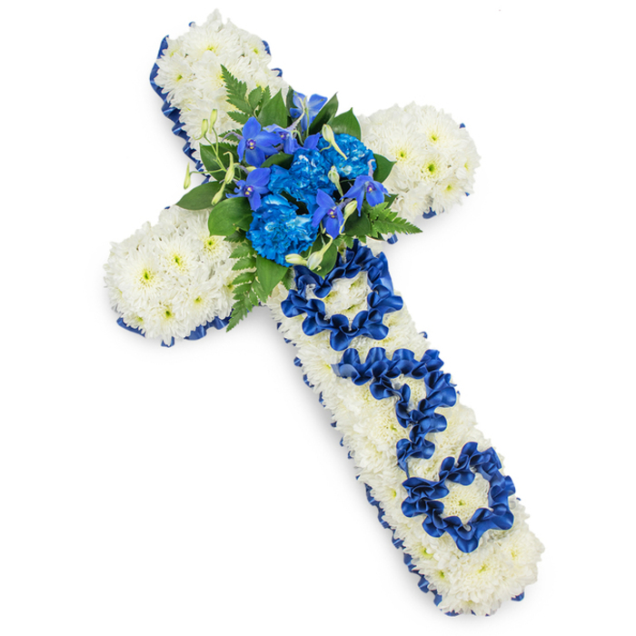 A detailed floral cross, clearly intended as a funeral or memorial tribute, rests on an immaculate white surface that accentuates its calm and reverent character. The entire cross is carpeted with white spray chrysanthemums placed so closely together that they create a smooth, pillowy texture, suggesting comfort and peace. Each small blossom is crisp and fresh, with soft petals that catch the gentle lighting, giving subtle shadows and depth across the surface. Framing this field of white, a royal blue satin ribbon runs along every edge, hugging the contours of the arms and vertical stem. The ribbon's rich colour and gentle sheen act like a frame around a painting, sharpening the outline of the cross and introducing a dignified, almost ceremonial feel. At the centre point, where the arms of the cross meet, a compact yet lush cluster of royal blue flowers rises slightly above the white background. Ruffled blue carnations are the stars here, their layered petals creating fullness, while more slender blue florets reminiscent of delphiniums or orchids add height and delicate detail. Deep green fern fronds and darker leaves encircle this central spray, adding contrast, texture, and a hint of garden freshness. Running down the length of the vertical arm, the word DAD is spelled out in ribbon, each letter shaped from loops and folds of royal blue satin, anchored into the chrysanthemum base with meticulous care. The effect is of a tribute that is both formal and deeply personal. This is the kind of thoughtfully made arrangement that might be delivered in Stanmore for a funeral at a local church or cemetery, offering a serene, faith-filled farewell to a much-loved father and reflecting the quiet strength and devotion of the family who sent it.