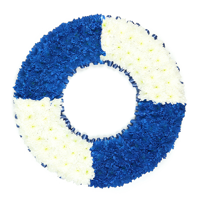 The photograph shows a refined sympathy wreath composed in the recognisable form of a lifebuoy, captured on a pure white backdrop that heightens its sense of calm and clarity. The ring is thoroughly covered with scores of small, tightly packed blooms, likely chrysanthemums, creating a dense, velvety surface where each little flower contributes to a greater whole. The design is split into four equal sectors that alternate in colour, moving blue-white-blue-white in a continuous sequence around the circle. The royal blue quarters at the top left and bottom right are particularly striking, with their deep, saturated tone suggesting the steadfast strength of the sea and perhaps echoing the uniform colours of naval or lifeguard service. In contrast, the white quarters at the top right and bottom left offer brightness and purity, aligning with the feelings of respect, remembrance, and peace that families in Stanmore often seek in a funeral tribute. Every petal is carefully placed, giving the wreath a polished, almost sculpted appearance. Along the inner circumference, a narrow blue ribbon has been delicately pleated into a fine ruffle, tracing the circle and framing the central hole, which reinforces the life ring symbolism. There are no added bows or greenery; the focus remains entirely on the disciplined pattern of blue and white and the associations it carries. As a memorial piece, this lifebuoy wreath feels especially suited to honouring someone with maritime ties, a fondness for the coast, or a role as a guiding figure in their family. Displayed at a church, crematorium, or graveside in Stanmore, it would stand as a dignified symbol of safe harbour, protection, and an enduring bond of love.