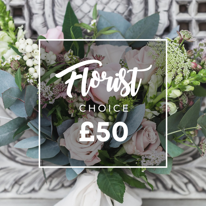 A lush, hand-tied floral arrangement fills the frame, centred on a cluster of soft blush-pink roses whose gently unfurling petals create a romantic, tactile focal point. Interspersed are bright white berries that catch light and bring lively contrast to the composition, while rounded, dusty-blue eucalyptus leaves form a soothing, silvery-green halo that cools and grounds the bouquet. Delicate sprays of tiny white filler blooms - likely waxflower or astrantia - float between the larger forms, giving an airy, natural cadence, and darker pink or burgundy accent blooms provide shadowed pockets of richness. Taller, slender green-and-white spikes rise at the left and right edges, adding a vertical grace reminiscent of snapdragons and introducing movement to the otherwise rounded bouquet. The stems are gathered into a neat hand-tied base and wrapped with a cream satin ribbon, visible at the bottom, signifying a bespoke finish. The bouquet is photographed against a softly blurred background of ornate carved stone or painted plaster and placed on an antique cream console, an elegant setting that lets the floral tones dominate the scene. In soft ambient light the arrangement reads as both soothing and enlivening - a perfect choice for a milestone birthday, a heartfelt thank-you, or as a refined housewarming gift. As a Stanmore florist I imagine this being delivered across local streets near Stanmore Common and Canons Park, chosen by customers who want fresh, seasonal blooms assembled with care and a quiet sense of luxury.