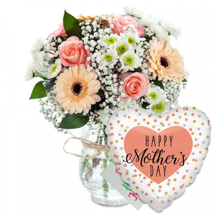 A beautifully composed Mother's Day scene is captured here, with all attention focused on a fresh bouquet and a celebratory balloon set against a crisp white background that enhances every colour and contour. On the left, a rounded bouquet of mixed spring flowers sits in a straightforward, jar-style clear glass vase. The stems inside are visible and neatly arranged, while a piece of light brown twine or raffia is tied casually around the neck, lending an informal, rustic charm that feels in tune with the green open spaces around Stanmore Common. The bouquet is a gentle blend of warm and cool pastels, designed to convey love without overpowering. Soft peach and pale pink roses are prominently placed, their petals opening in generous layers and curling back at the edges like waves, each flower embodying tenderness and appreciation. Two large peach gerbera daisies with sharply defined dark brown centres break through the softness with a dose of bright, uncomplicated happiness, their flat, circular faces anchoring the composition. Throughout the bouquet, fine clusters of white baby's breath weave in and out, forming tiny stars that lend lightness and delicacy. Smaller white chrysanthemum blooms, each with a hint of pale green at the centre, add density and a crisp, clean tone, filling the arrangement so that it feels abundant yet still airy. Glossy green foliage frames the blooms and extends just beyond their edges, providing depth, contrast, and the sense of flowers freshly chosen from a local Stanmore florist. To the lower right, partially obscuring the vase, a shiny heart-shaped foil balloon brings a touch of playful glamour. The balloon's base colour is bright white, covered with scattered gold and silver polka dots that look like celebratory confetti. In the centre, a solid peach or light coral heart contains the printed message HAPPY Mother's DAY, where HAPPY appears in bold, straightforward lettering and Mother's DAY is rendered in an elegant script. Between the balloon and the vase, the corner of a small white greeting card peeks through, hinting at a handwritten message. The whole image evokes a complete, carefully considered Mother's Day gift, perfect for delivery to homes and family gatherings across Stanmore.