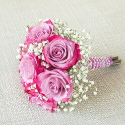 Bathed in gentle natural light, this beautifully composed bouquet channels the romance of a Stanmore celebration, its vibrant colour palette a harmonious blend of tradition and modernity. Five large roses, their tight, velvety petals blushing from soft lavender at the centers to electrifying fuchsia around the outermost curves, serve as the focal point. Among them, airy tufts of tiny white baby's breath blossoms dance, creating a soft, cloud-like atmosphere and offering elegant contrast to the saturated pinks and purples. The stems are gathered in a careful, compact arrangement and wrapped with deep purple satin ribbon-its texture both tactile and visually rich-accented all round by meticulously placed, spiraling rows of gleaming pearls, reminiscent of bridal jewelry. The green and brown ends of each stem appear at the bottom, evoking a sense of freshness and handpicked authenticity, perhaps gathered in the early hours from local nurseries near Stanmore Park. The bouquet lays atop a neutral, textured surface, its colours popping against the subtle linen, much like floral displays seen at Stanmore's celebrated events. Every detail-colour, texture, and style-suggests both innocence and sophistication, making this bouquet a fitting companion for a Stanmore registry wedding, an elegant bridal shower, or a cherished family celebration by the leafy avenues of HA7.