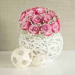 A delicate, romantic still life captures a domed bouquet of pink roses and baby's breath arranged with the precision and softness you might expect from a trusted Stanmore florist. The roses are tightly grouped, forming a near-perfect sphere, their petals unfurling from creamy, blush-pink centres into richer pink tips that border on fuchsia. Between the roses, airy clusters of white gypsophila weave in and out, creating the impression of tiny clouds resting among the blooms and lending a sense of freshness, like a cool morning stroll through a garden off Marsh Lane. The arrangement is cradled in a round white vase made from intricately woven natural fibres, the open latticework pattern letting glimpses of shadow and light interplay across the smooth white surface beneath. To one side, two decorative spheres crafted from pale wooden or rattan strips continue the theme of woven texture, their simple forms echoing the vase while keeping the focus firmly on the flowers. The background is a softly textured wall in warm beige or cream, gently illuminated so that no harsh shadows disturb the calm, inviting feel. The overall effect is one of understated elegance and contemporary charm, perfect for brightening a dining table in a Stanmore townhouse, welcoming guests in a hallway, or marking sentimental moments such as anniversaries or new beginnings. The arrangement seems ready for same-day delivery across Stanmore, capturing that blend of modern styling and timeless affection that suits both everyday gestures and special celebrations.