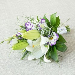 Resting diagonally on a pale, textured beige cloth that resembles finely woven linen, this hand-tied bouquet feels like a moment of quiet luxury captured in time. The eye is first drawn to the open white Oriental lilies, their broad, velvety petals folding back gracefully to reveal deep yellow-orange stamens and subtle maroon speckling, each flower like a star unfolding on the fabric. Alongside them, several plump lime-green buds lean outwards, promising that the bouquet will keep changing and opening over the days ahead. Interlaced among the lilies, Lisianthus blooms introduce a playful contrast: white, ruffled petals with bold purple rims and centres that wash from lilac to rich violet, like brushstrokes of colour. Tiny sprays of white Gypsophila weave through the arrangement, their minuscule blossoms forming a light mist that softens the strong forms of the lilies and Lisianthus. Surrounding everything, layered greenery in a range of tones and textures-polished dark green leaves, fine wisps, and slightly spiky accents-creates a lush, natural frame and a gentle sense of movement, as though a breeze has just passed through. The stems are carefully gathered and wrapped with a smooth white ribbon, where delicate pearl-like beads glint subtly along the narrow, right-hand taper, giving a nod to bridal jewellery and special occasions. The lighting is soft and even, with no harsh glare, allowing the whites to look creamy rather than stark and the purples to remain rich. It is easy to imagine this bouquet being carried from a ceremony in one of Stanmore's churches, or arriving as a same-day delivery surprise to a home near Stanmore Country Park, crafted with the calm confidence of an experienced local florist.