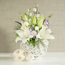 This image shows a refined floral centrepiece of white lilies and lisianthus styled in a way that suits a Stanmore wedding breakfast or an elegant dinner party near Canons Park. The flowers are gathered in a distinctive round white vase with an open lattice, basket-like design, allowing glimpses of stems and light through its woven structure and giving an airy, modern feel. Several lilies dominate the composition, their large white blooms fully open to reveal soft green throats and pollen-tipped stamens, while a few tall, tapered green buds inject height and a sense of evolving life. Threaded between the lilies are lisianthus stems, some still curled into smooth buds and others displaying gently ruffled white petals brushed with delicate lavender along the rims, adding subtle colour and romance. Small sprays of white filler florets, similar to gypsophila, nestle among the main flowers, and occasional tiny clusters of deeper purple blossoms add quiet depth without overwhelming the restrained palette. Surrounding and supporting the blooms, varied greenery spills over the lip of the vase, combining fine, fern-like textures with broader, more structured leaves to create movement and softness. The arrangement is placed on a sleek, bright white surface, flanked by two woven decorative spheres made from light beige material; their open, ribbon-like strands echo the vase's pattern and reinforce the natural, contemporary aesthetic. In the background, a light beige wall with a horizontal, linen-like texture is bathed in gentle daylight, enhancing the whites, greens, and lilac accents so they appear fresh and luminous. The overall effect is one of quiet celebration and comfort, making this design ideal for wedding table arrangements, sympathy tributes, or thoughtful gifts delivered to homes and venues throughout Stanmore and nearby areas.